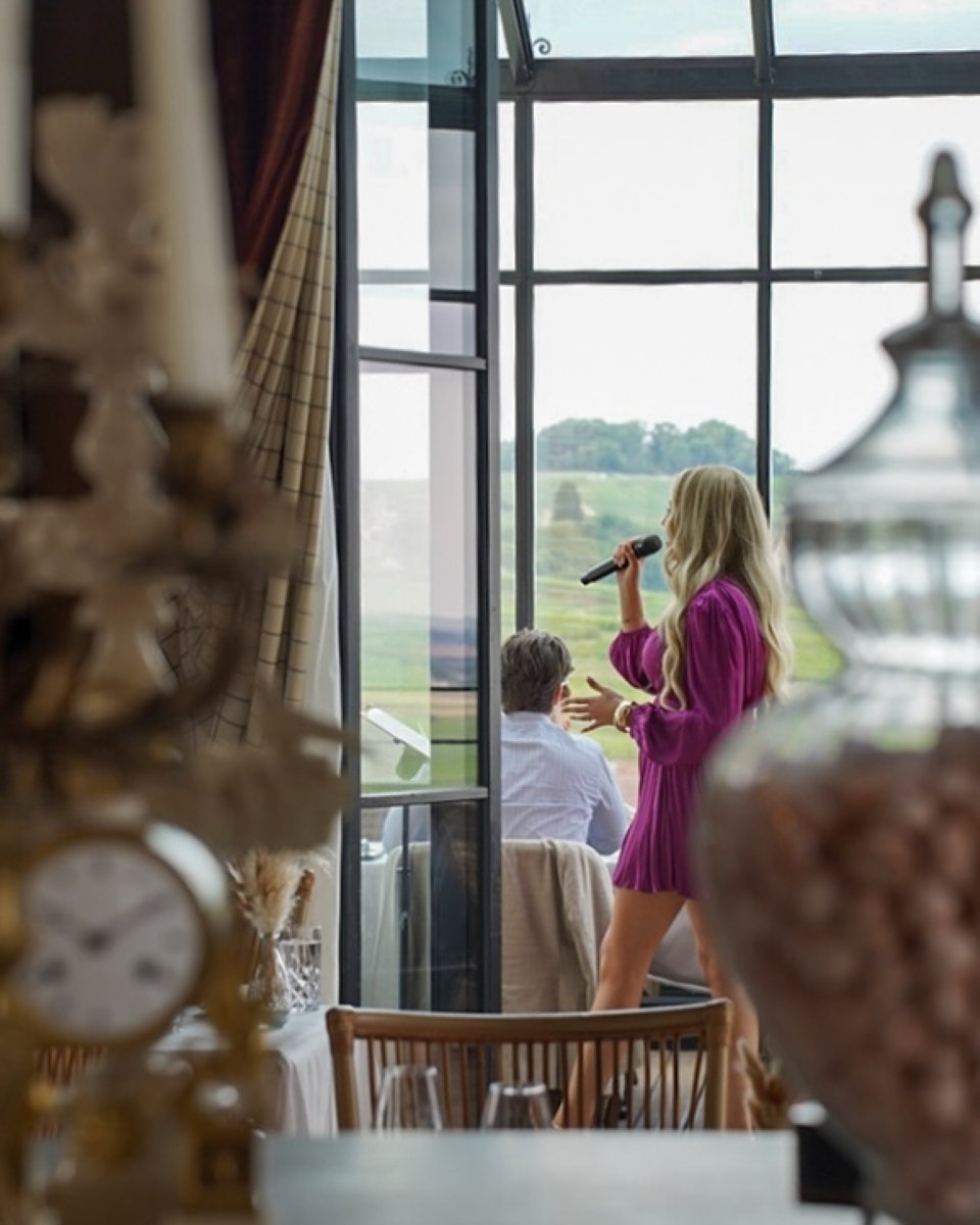 Zangeres Vonne zingt in een roze jurk bij een groot raam met uitzicht op het landschap