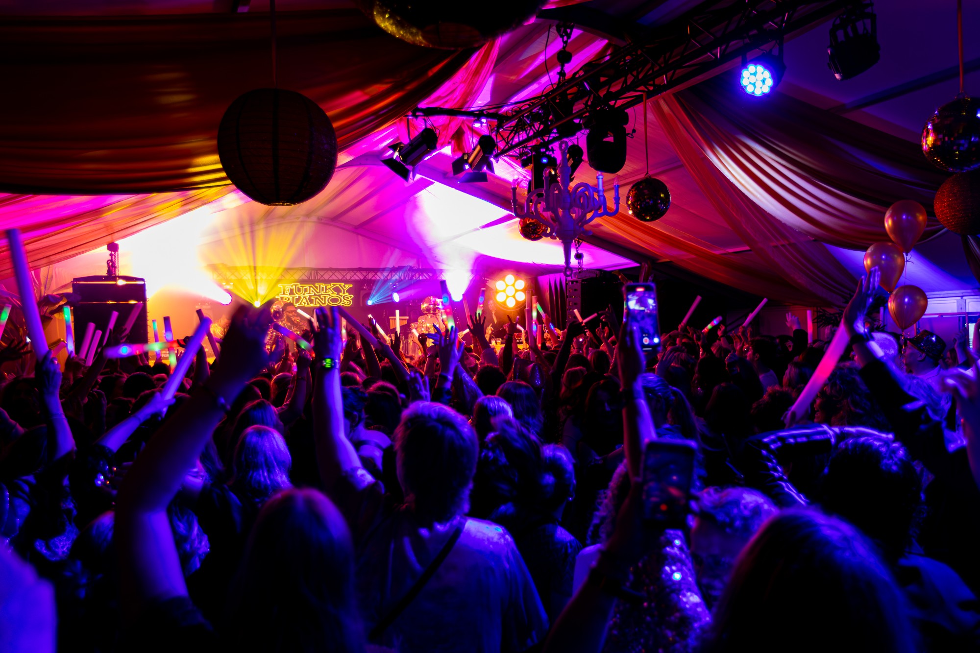 Dansende gasten met lichtgevende staafjes voor het podium van Funky Pianos in een feesttent