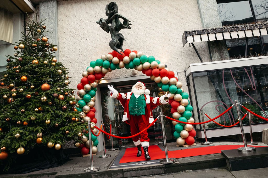 De kerstman staat onder een ballonnenboog met rode loper bij ’t Slaakhuys, naast een kerstboom