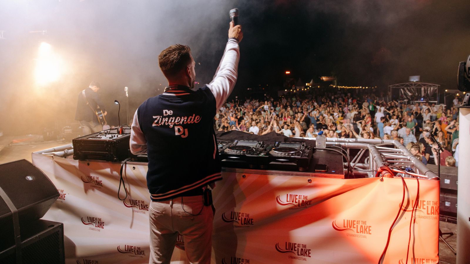 De Zingende DJ steekt zijn microfoon in de lucht voor een volle festivalweide